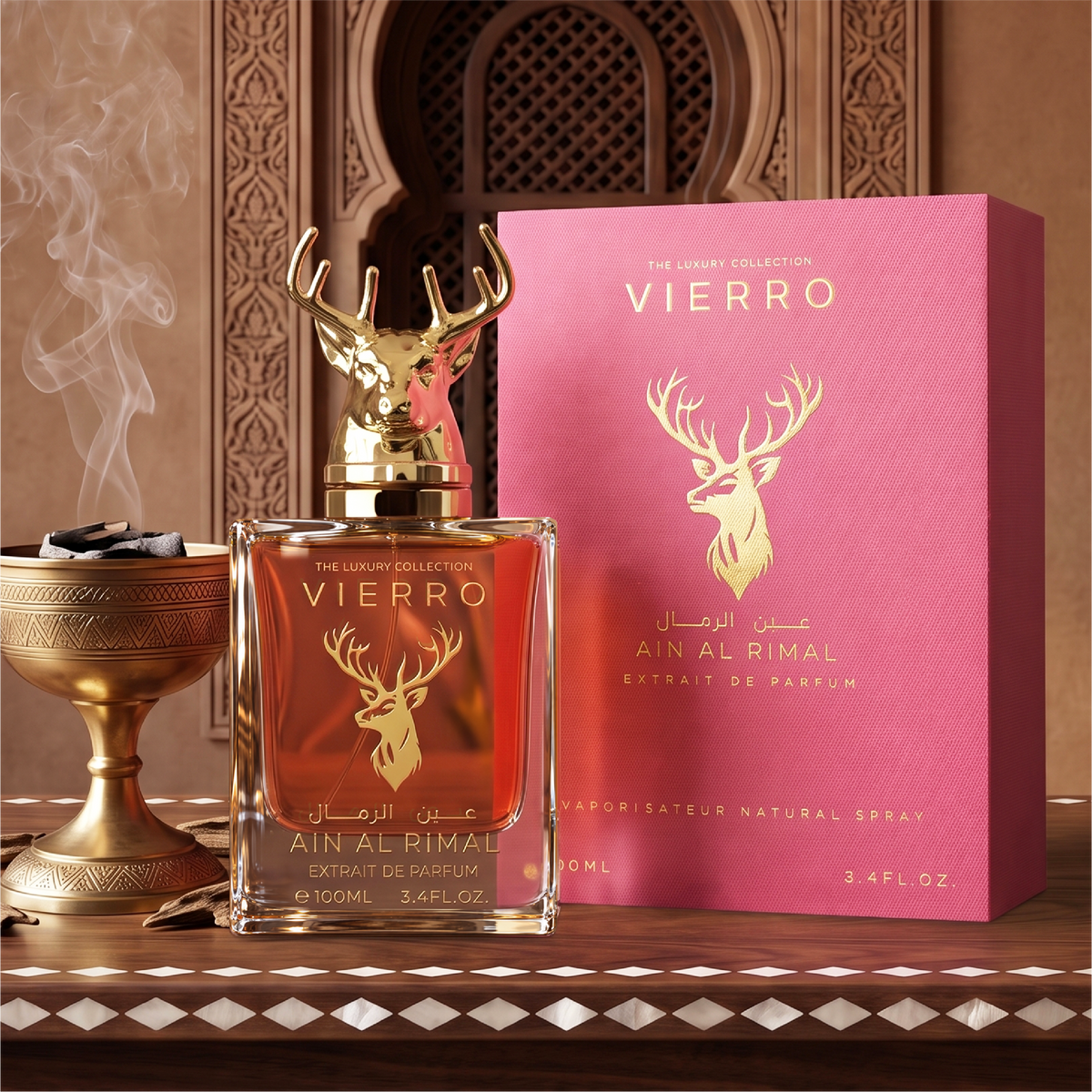 A 100ml bottle of Ain Al Rimal Extrait de Parfum by Vierro featuring a gold deer head cap and amber liquid, displayed next to its matching pink product box on an ornate wooden surface with an incense burner in the background.